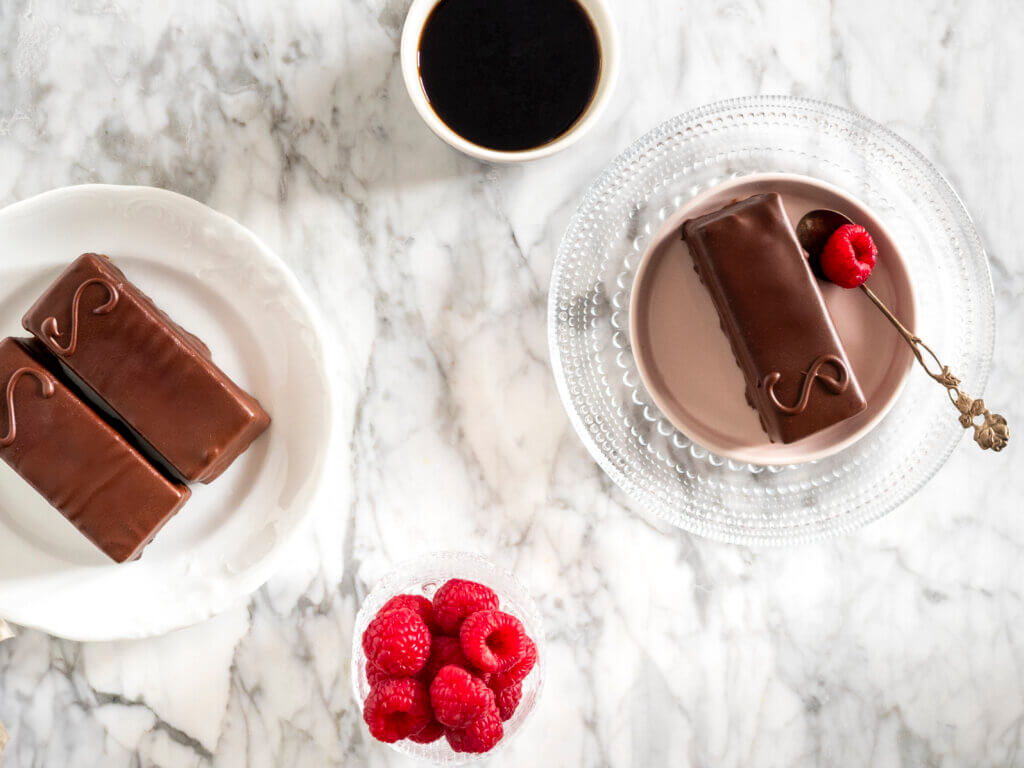 Marmoripöydällä kolme Sacherleivosta, kuppi kahvia ja vadelmia.